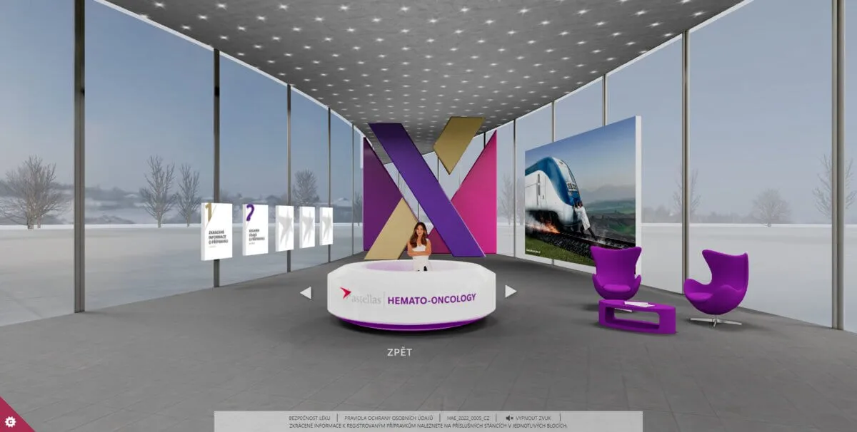 Hemato-onkologická recepce uvnitř 3D pavilonu Astellas: prosklená hala stojící ve sněhové pláni, natočená moderátorka za brandovým pultem, informační panely vznášející se ve vzduchu a za ní barevná socha oddělení.