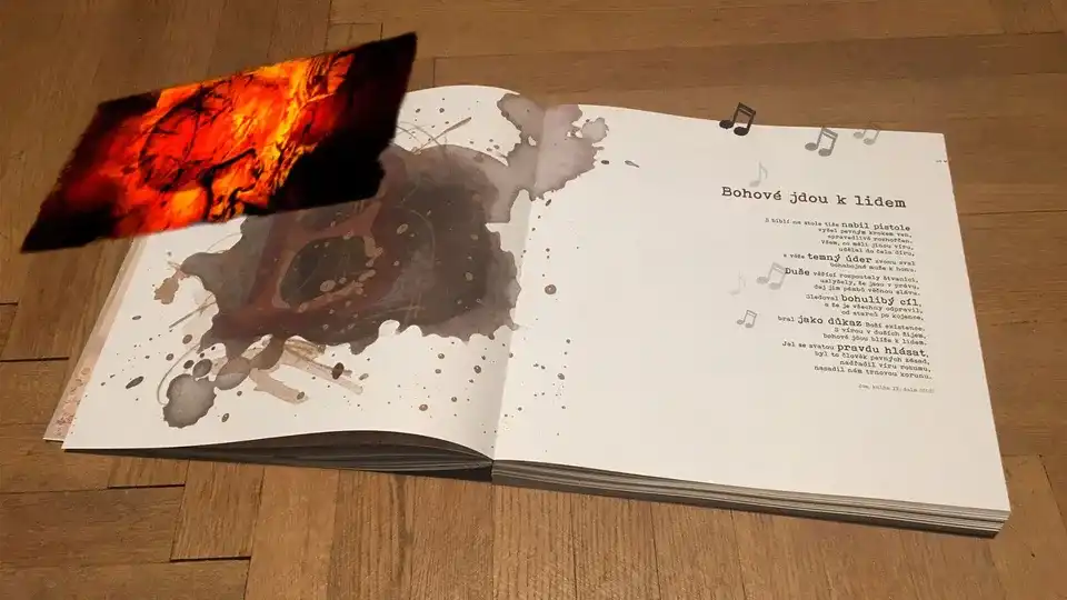 The book open on a wooden floor at the song “Bohové jdou k lidem”, ink blots across the left page — and a panel of burning embers standing in the air above it, played by the app.